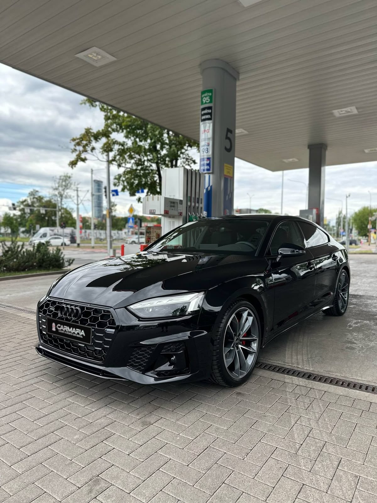 Czarne Audi A5 Sportback z przodu, pod wiatą stacji paliw, z tablicą CarMafia.fun