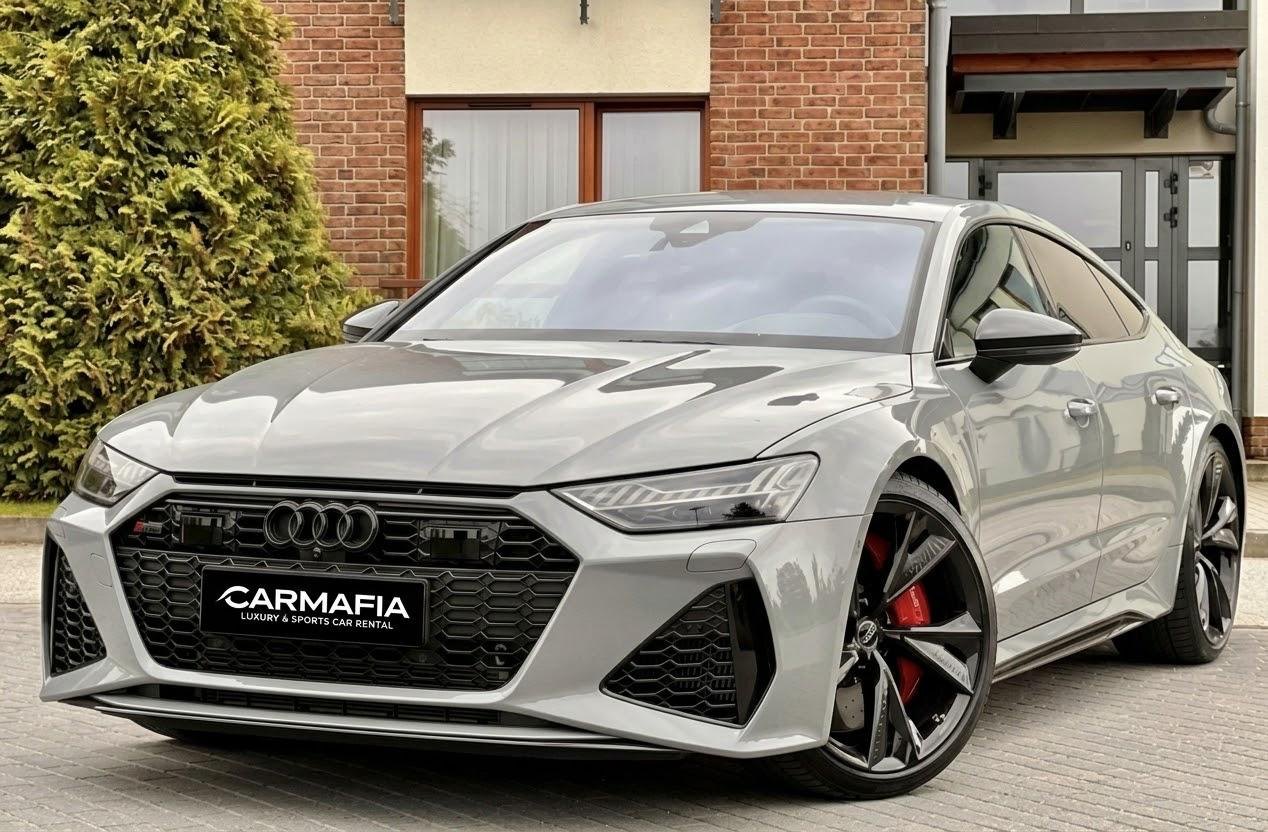 Grey Audi RS7 from the front at an angle, with red brake calipers and a CarMafia plate, in front of a brick house