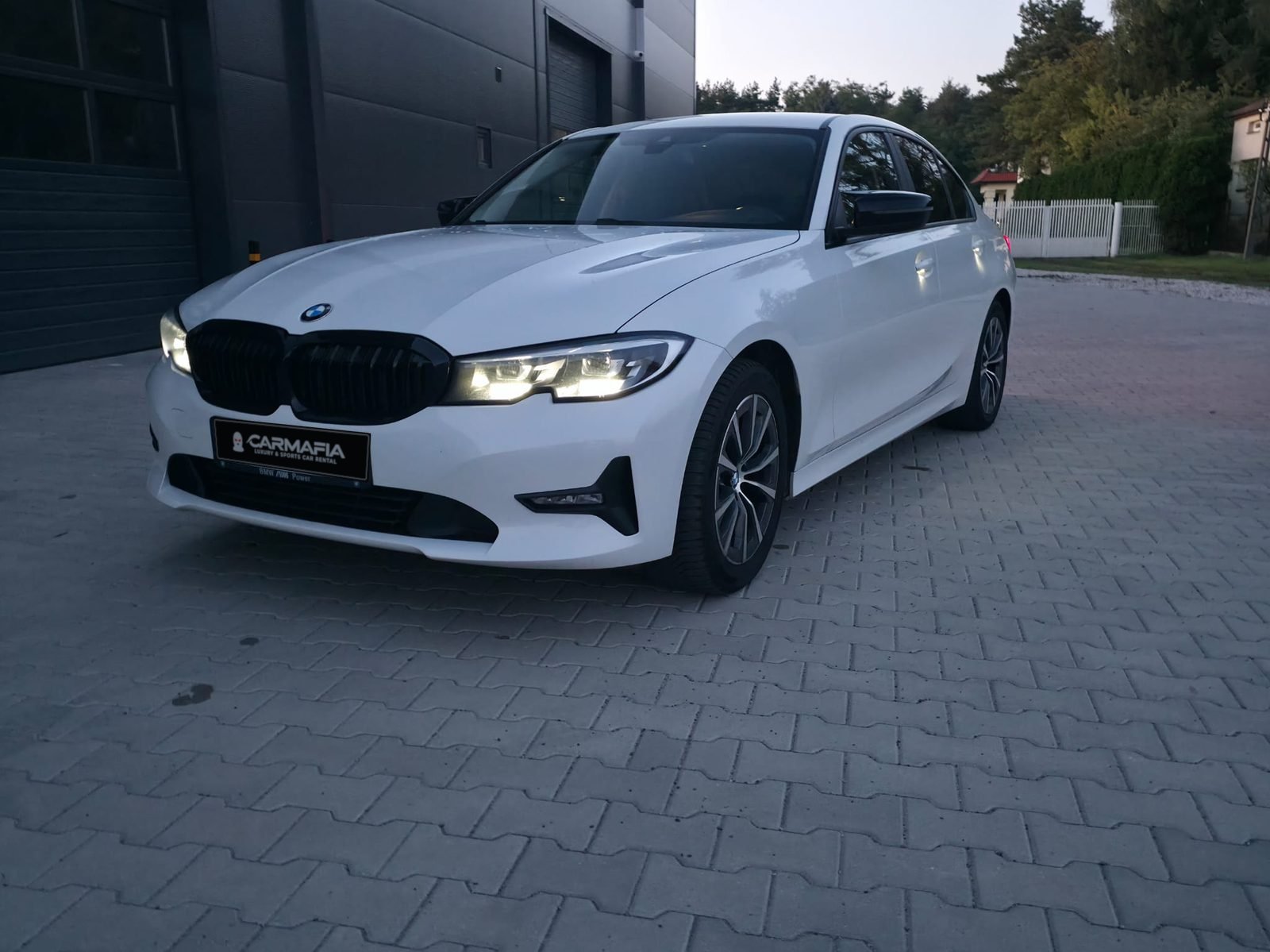 Białe BMW 318i G20 2020 z przodu, z włączonymi reflektorami, o zmierzchu