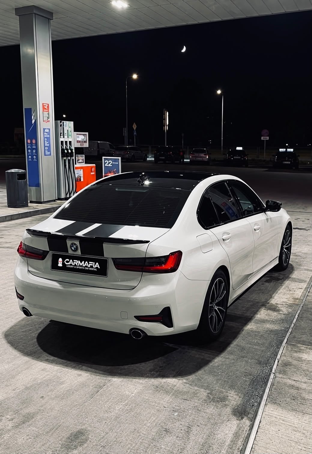 Białe BMW serii 3 z floty CarMafia.fun nocą na stacji paliw, widok z tyłu na tablicę z logo CarMafia.fun